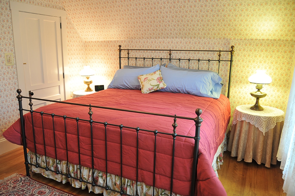 Elia Corti bedroom with King bed black metal bed frame , two round end tables with lace and brocade covers and period lamps. Flowered wall paper