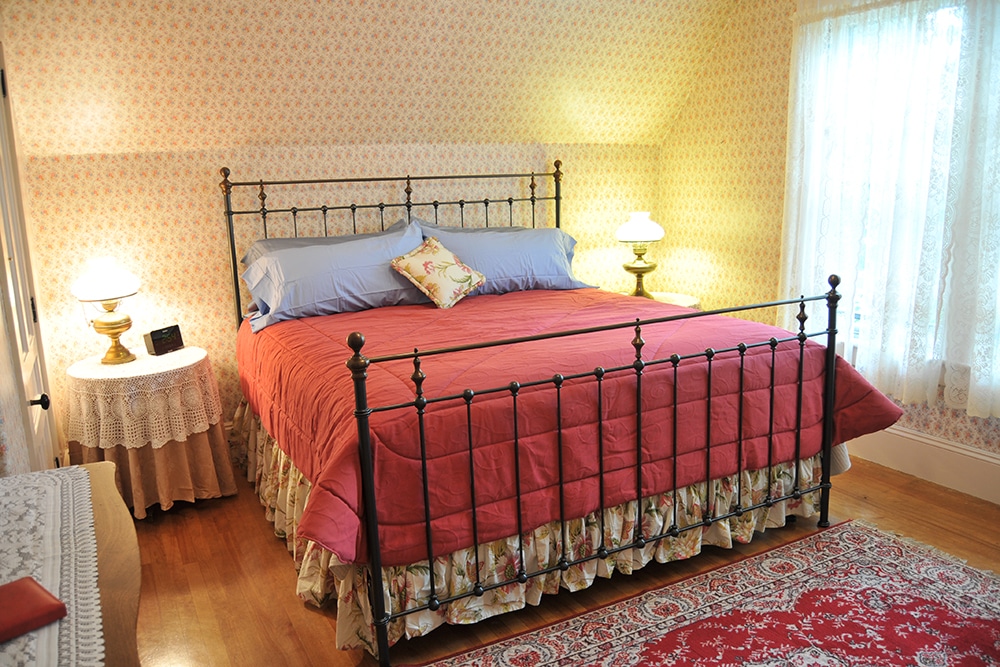 Elia Corti bedroom with King bed black metal bed frame , two round end tables with lace and brocade covers and period lamps. Flowered wall paper. Oriental rug predominantly red
