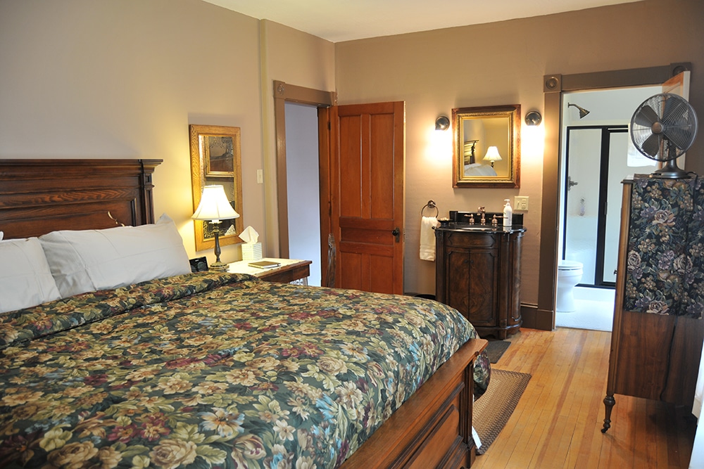 Large wooden headboard and footboard on generous king bed with floral patterned comforter. Wooden end table with lamp. Carved dark wood and marble vanity with white hand towel, hand soap and lotion on marble top. Gold framed mirror above. glimpse into newly renovated bathroom white tile and commode . Antique wooden dresser with black fan on top