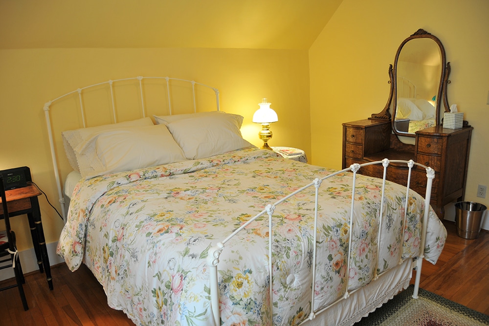 Yellow walls with queen bed. White metal head and foot boards with floral comforter. hard wood floor, antique writing desk and chair with antique lamp and round end table with antique lamp, ornate wood dresser.