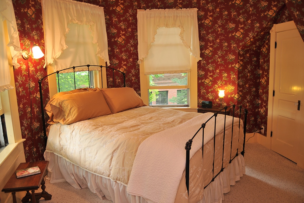 Round room with wallpaper red background with cream and green flowers beige, carpet. Queen bed with metal head and foot board. cream and white comforter with white throw blanket and bed skirt. Wood end tables with antique lamp
